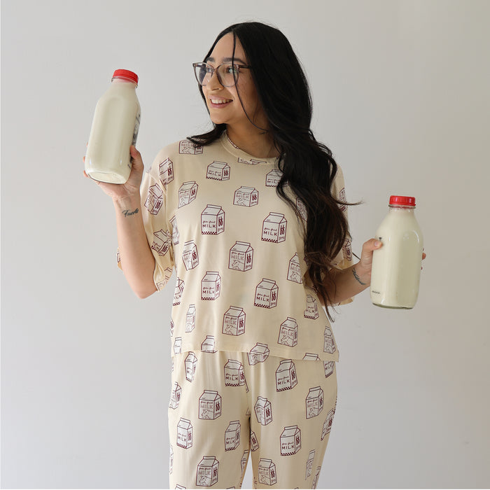 A smiling woman wears the forever french baby Women's Boxy Tee Lounge Set in "Milk" and holds two glass bottles of milk, standing against a plain white background.