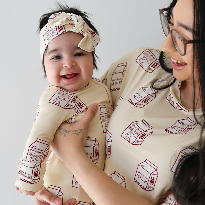 A smiling baby in a Forever French Baby outfit with red milk carton prints and a Bamboo Head Wrap | Milk, held by an adult in matching cream outfits—both sporting the charming look from forever french baby.