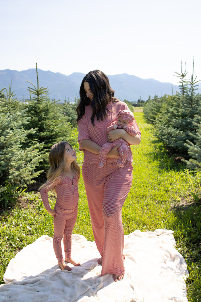 A woman holding a baby stands next to a young girl, all dressed in matching forever french baby Women's Boxy Tee Lounge Sets in Peppermint Stripe Ribbed, posing on a blanket among small evergreen trees with mountains visible in the background.