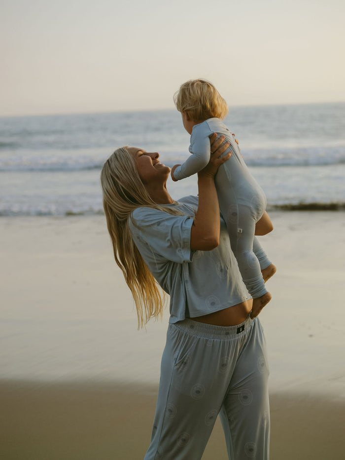 A woman in the Women's Boxy Tee Lounge Set | San Clemente by forever french baby lifts a toddler in matching pajamas on a sandy beach at sunset, with ocean waves in the background.