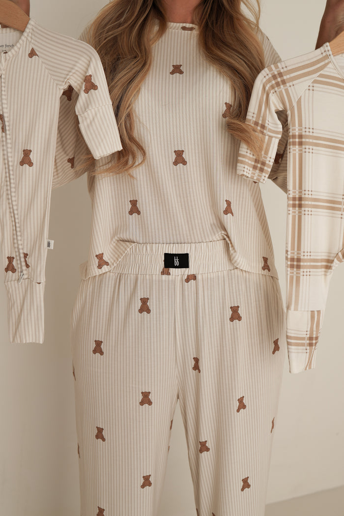 Wearing the forever french baby Women's Boxy Tee Lounge Set | Teddy, a person holds up two matching children's pajamas, one in each hand. Their cream-colored bamboo set features brown teddy bear prints. The person's face is not visible.