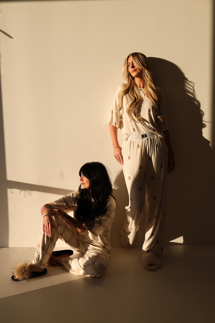 Two women wear the Women's Boxy Tee Lounge Set | Teddy by forever french baby, posing against a cream wall in warm sunlight. Made from hypo-allergenic bamboo, these comfy outfits radiate relaxation—one standing with hands in pockets, one sitting cross-legged.