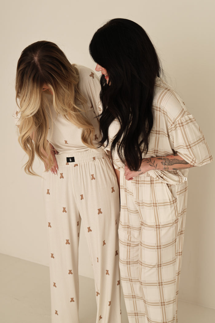 Two women smile together, wearing the forever french baby Women's Boxy Tee Lounge Set | Teddy in light tones—one in plaid, the other with brown shapes—against a plain, light background.