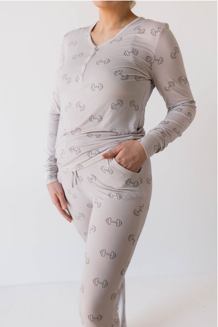 A woman is wearing a grey, long-sleeve top and matching pants adorned with a pattern of small black dumbbells. The stylish outfit looks like it could be part of the trendy Women's Bamboo Pajamas | Arm Day collection from forever french baby. She stands with one hand in her pocket against a plain white background.