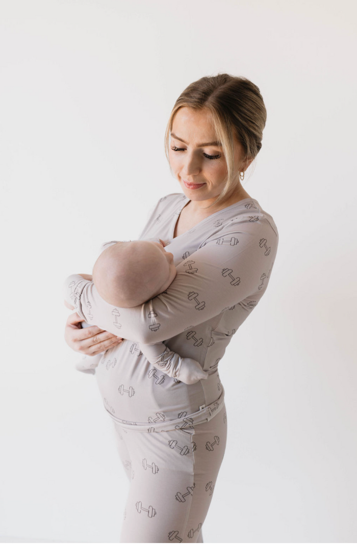 A woman with light hair is gently holding a baby in her arms. They are both dressed in matching light-colored Women's Bamboo Pajamas | Arm Day by forever french baby, featuring dumbbell weight designs. The background is plain white. The woman looks down at the baby with a tender expression.