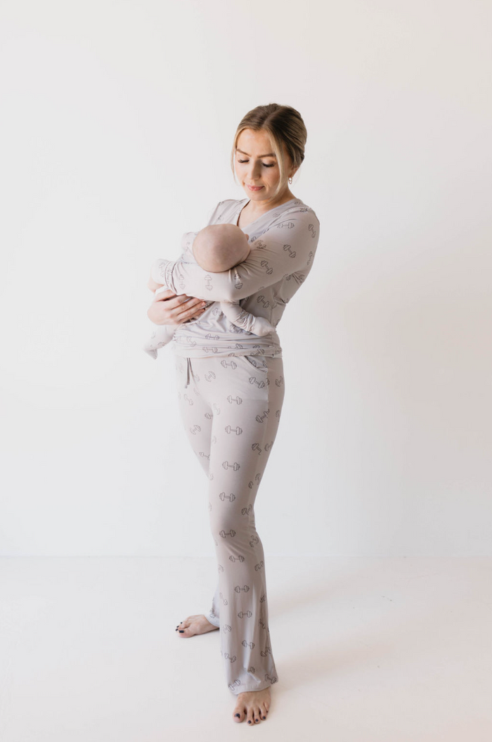 A woman stands against a plain white background, holding a baby in her arms. Both are wearing matching light gray outfits adorned with small dumbbell weight designs. The woman, dressed in forever french baby's Women's Bamboo Pajamas | Arm Day, gazes at the baby with a gentle expression.