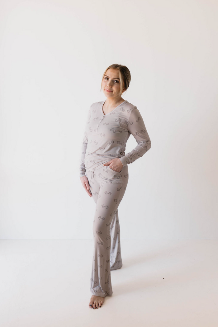 A woman in light gray pajamas adorned with a subtle pattern stands barefoot against a plain white backdrop. She is smiling, with one hand resting on her hip and the other tucked into the pocket of her forever french baby Women's Bamboo Pajamas | Arm Day, showcasing an effortlessly stylish and eco-friendly choice.