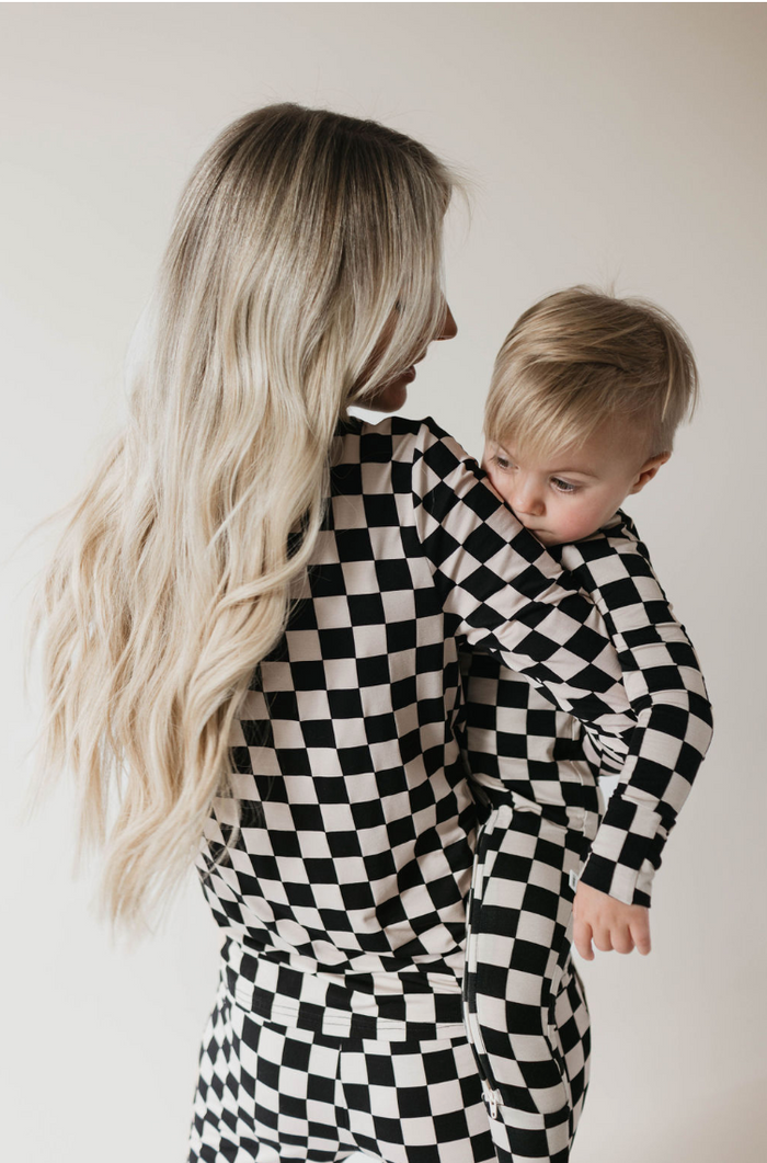 A woman with long blonde hair, clad in Forever French Baby's Women's Bamboo Pajamas in Black Checkerboard, holds a young child dressed in matching bamboo fabric pajamas. Both are facing slightly away from the camera against a light background.