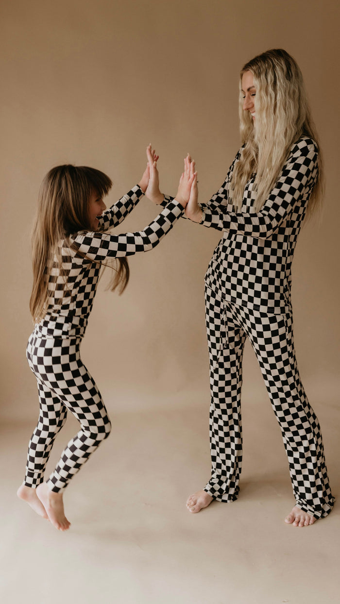 In a neutral-toned room, a young girl and a woman with long hair, both dressed in forever french baby Women's Bamboo Pajamas in Black Checkerboard, playfully touch hands. The girl stands on one leg, and both are smiling as they share a fun moment.