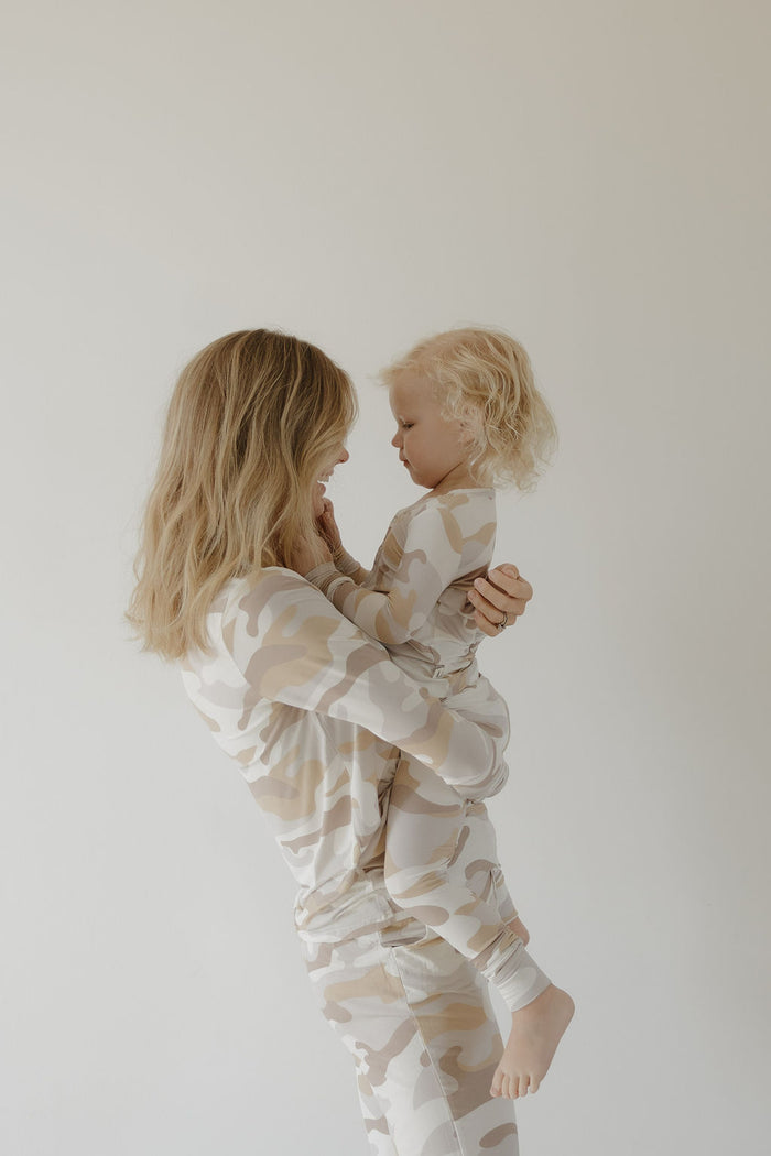 A woman holds a young child in her arms, both wearing forever french baby Bamboo Two Piece Pajamas | Camo 2.0, sharing a tender moment together against a plain, light background.