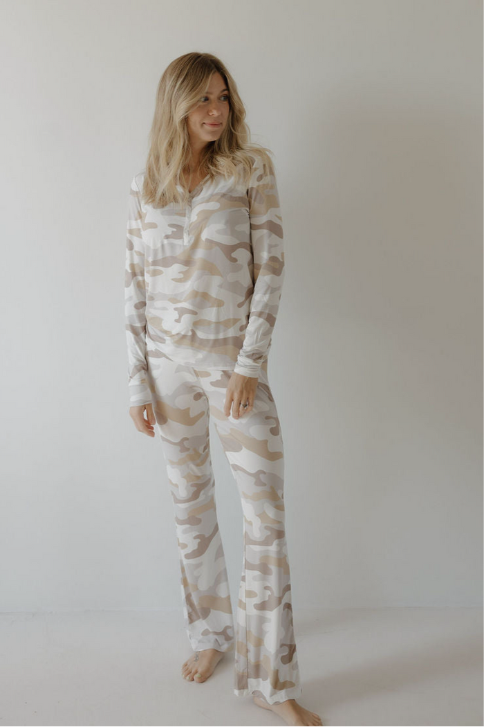 A woman with light brown hair wears forever french baby’s Women's Bamboo Pajama | Camo 2.0—long-sleeve, beige and white camo print. She stands barefoot against a white wall, softly smiling. The breathable bamboo fabric ensures all-night comfort.