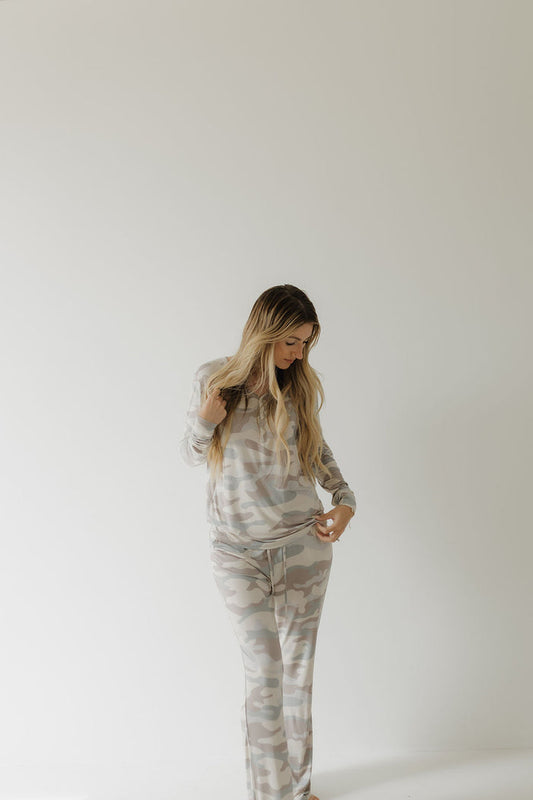 A person with long blonde hair wears the Women's Bamboo Pajama in camouflage by forever french baby. Against a plain white background, they look downward while slightly adjusting their hypoallergenic gray and white outfit.