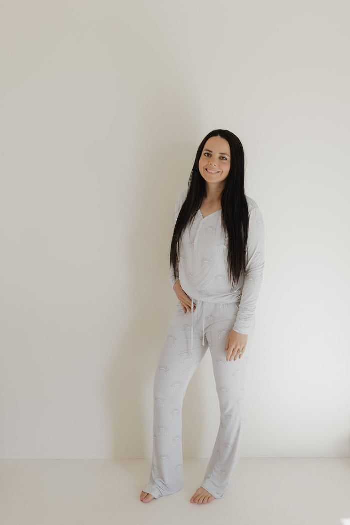 A person with long dark hair stands barefoot against an off-white wall, wearing forever french baby's Women's Bamboo Pajama | Howdy. They look ahead with a relaxed expression and hands in pockets, embodying the tranquility of breathable clothing.