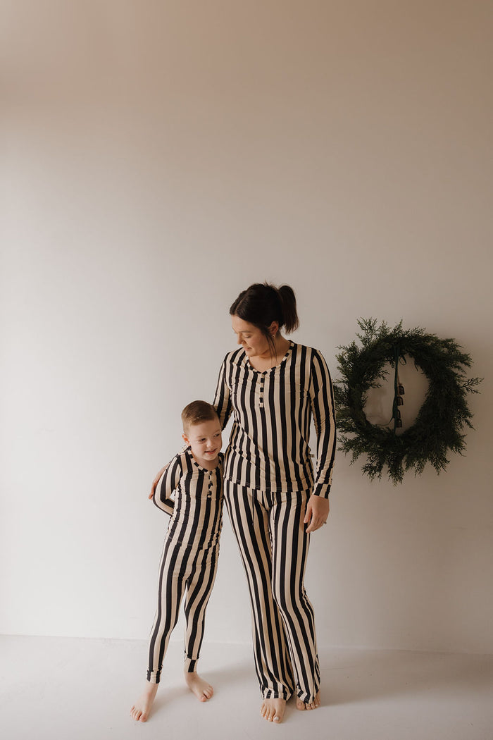 A woman and child stand barefoot in matching forever french baby Women's Long Sleeve Bamboo Pajamas in Midnight Stripe, with the woman’s arm around the child and a green wreath hanging on the beige wall behind them.