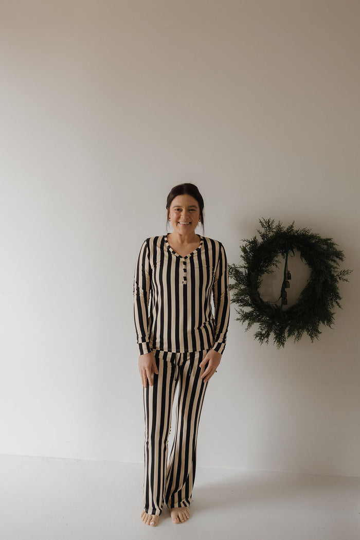 A woman smiles at the camera barefoot indoors, wearing the forever french baby Women's Long Sleeve Bamboo Pajama in Midnight Stripe. A green wreath on a white wall highlights her stylish and breathable bamboo sleepwear.