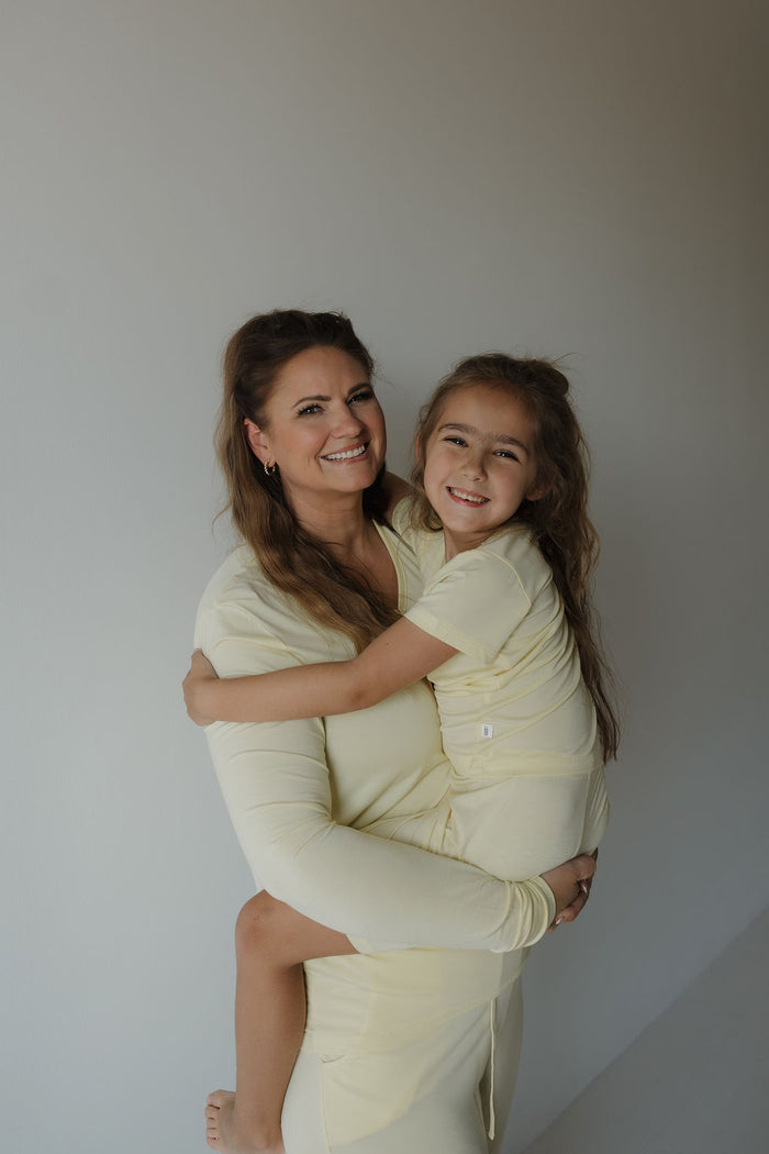 A woman wears the forever french baby Women’s Long Sleeve Bamboo Pajama in Sunny, holding a smiling young girl in matching yellow attire. Both face the camera against a plain light-colored background, highlighting the comfort and style of their hypo-allergenic bamboo clothing.