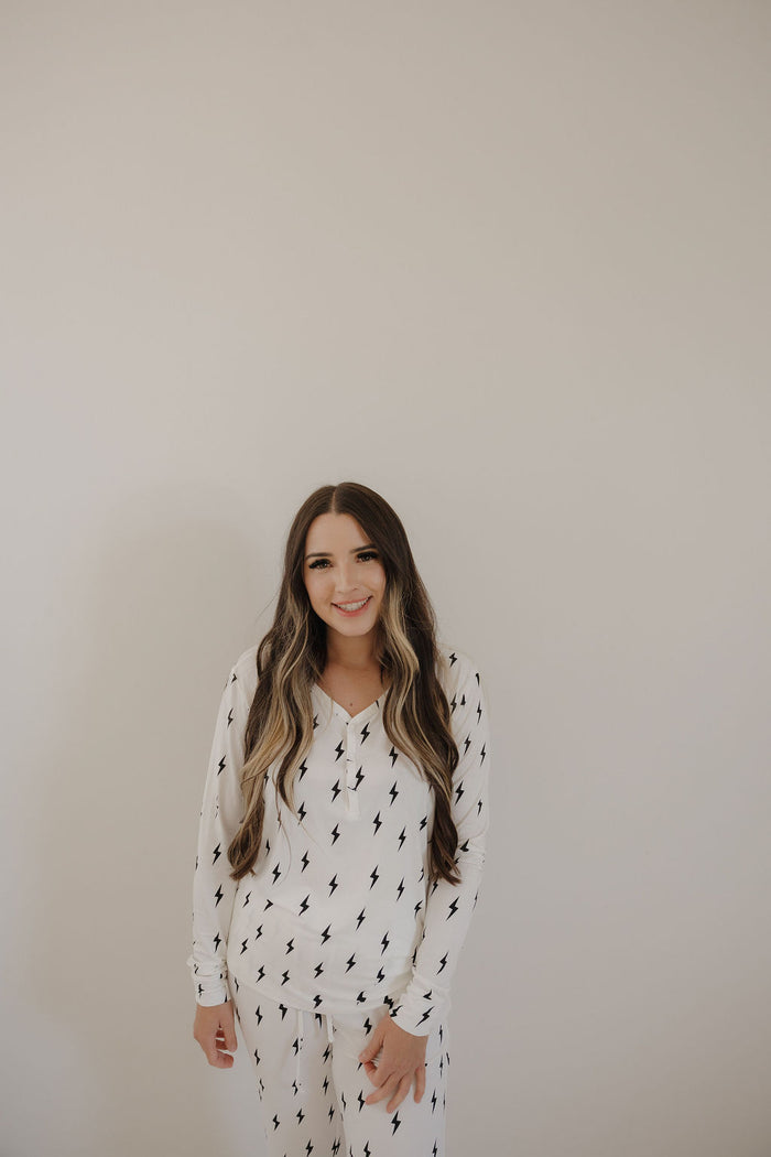 A woman with long, wavy brown hair smiles against a light background in forever french baby’s Women’s Bamboo Pajama | White & Black Lightning Bolt, featuring a breastfeeding-friendly design and black lightning bolt print.