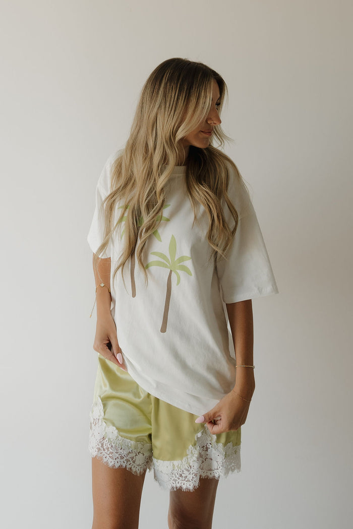 A woman with long, wavy hair wears a relaxed fit white T-shirt featuring palm tree graphics and light green forever french baby Women’s Satin Shorts | Prickly Pear, trimmed with lace, standing against a plain, light background.