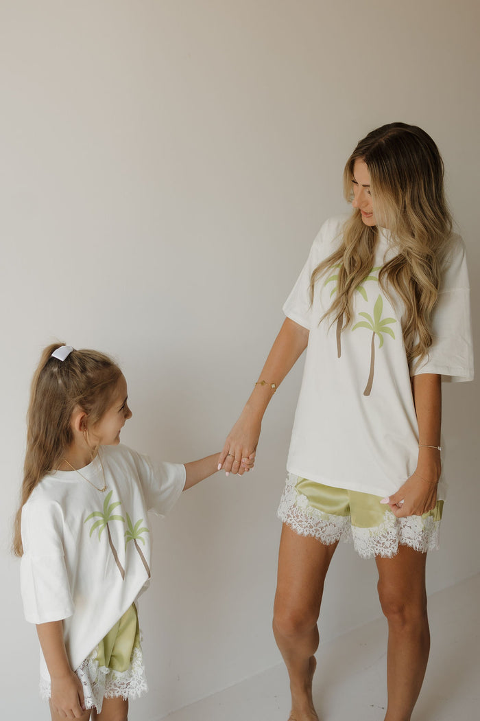 A woman and a young girl hold hands, both wearing matching forever french baby Women’s Satin Shorts in the “Prickly Pear” shade and white shirts with palm tree prints. Their relaxed fit green satin shorts add a playful touch as they stand against a plain light background.